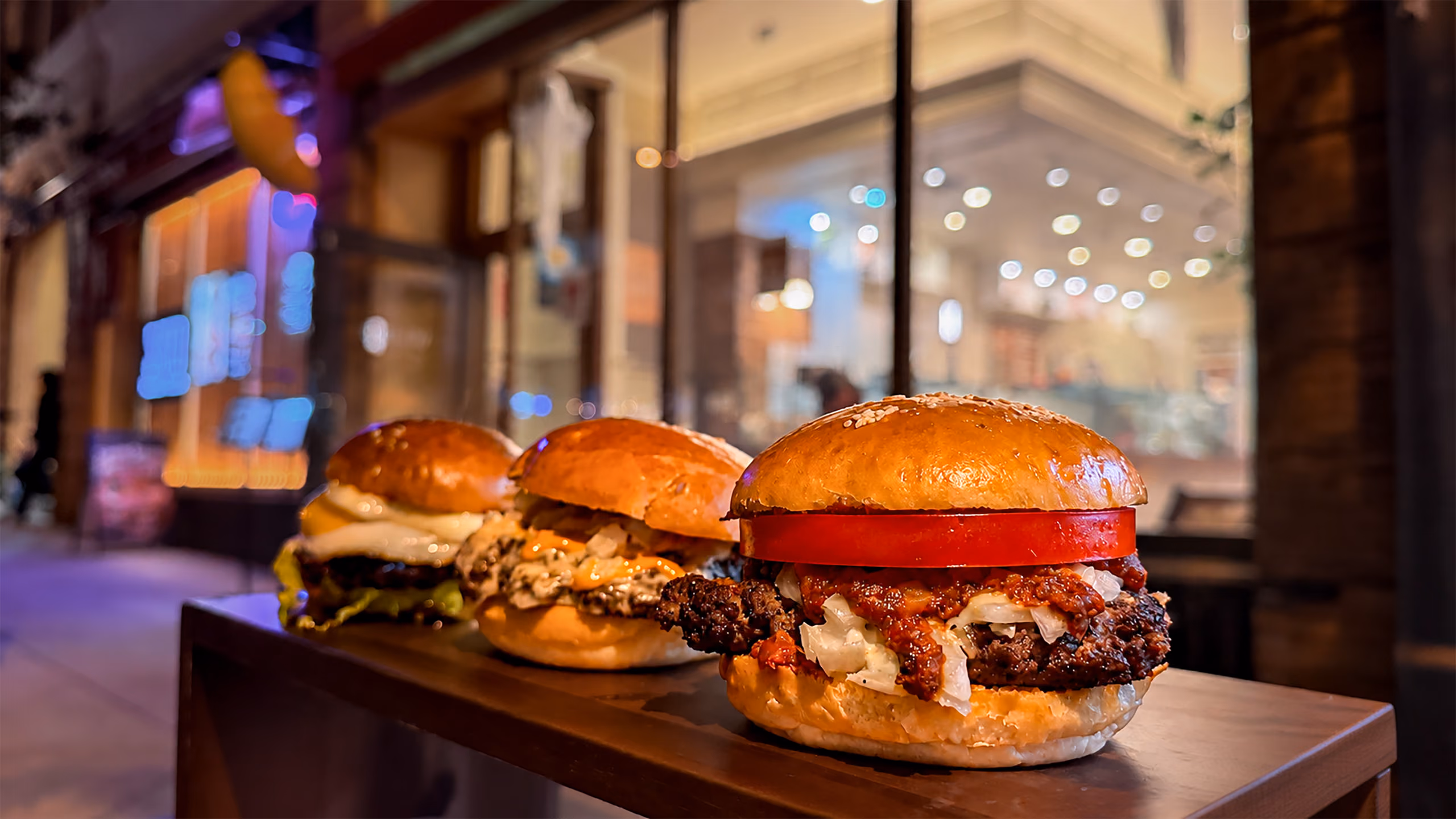 Three burgers on sesame brioche buns lined up on a wooden ledge outside the café at night, the nearest topped with a thick tomato slice over a chunky meat sauce, the lit storefront blurred behind them.
