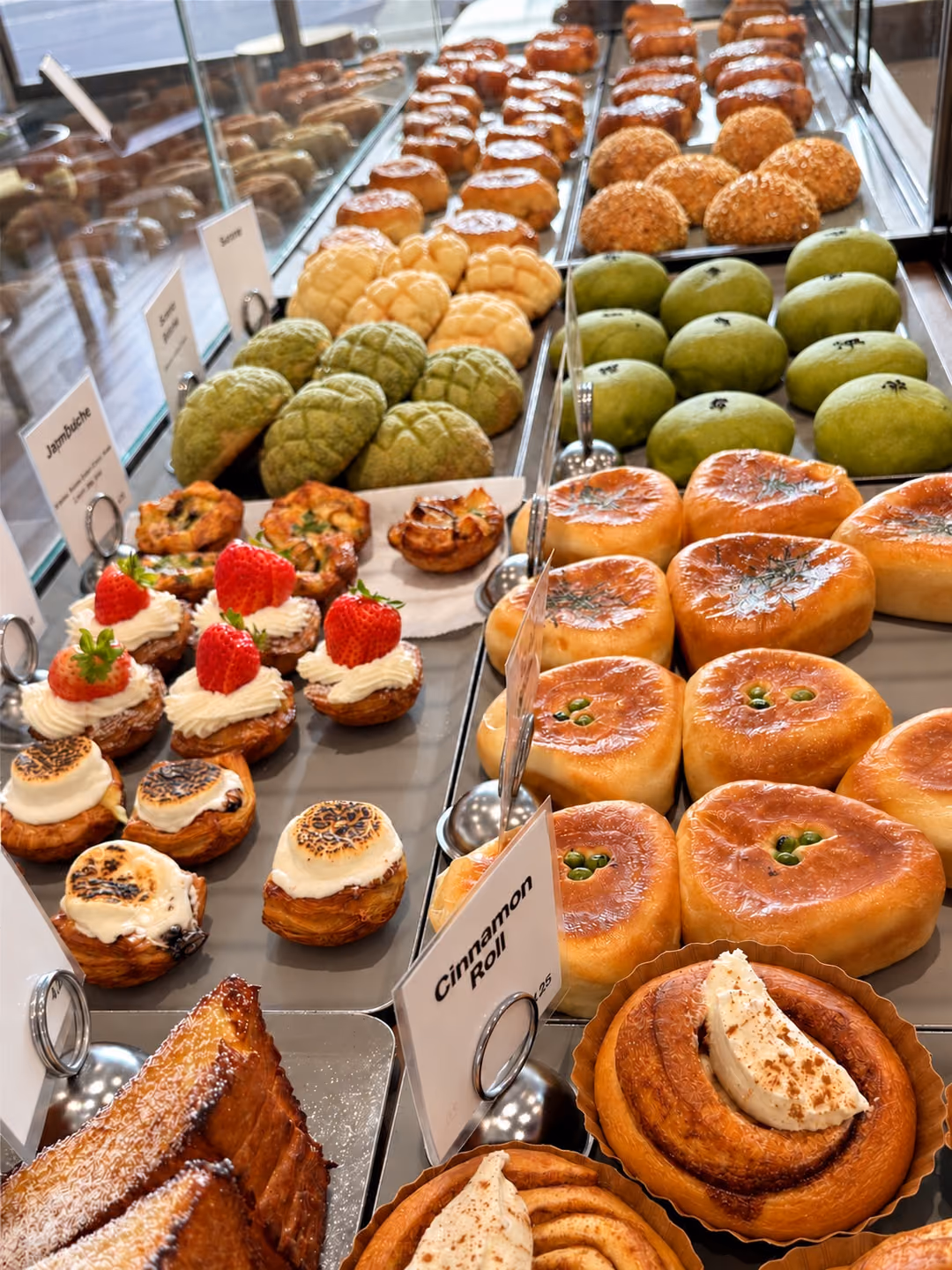 A bakery case seen down its length — cream-topped cinnamon rolls and sugar-dusted French toast in front, then soft buns, strawberry- and marshmallow-topped danishes, matcha and plain melon pan, and more trays beyond.